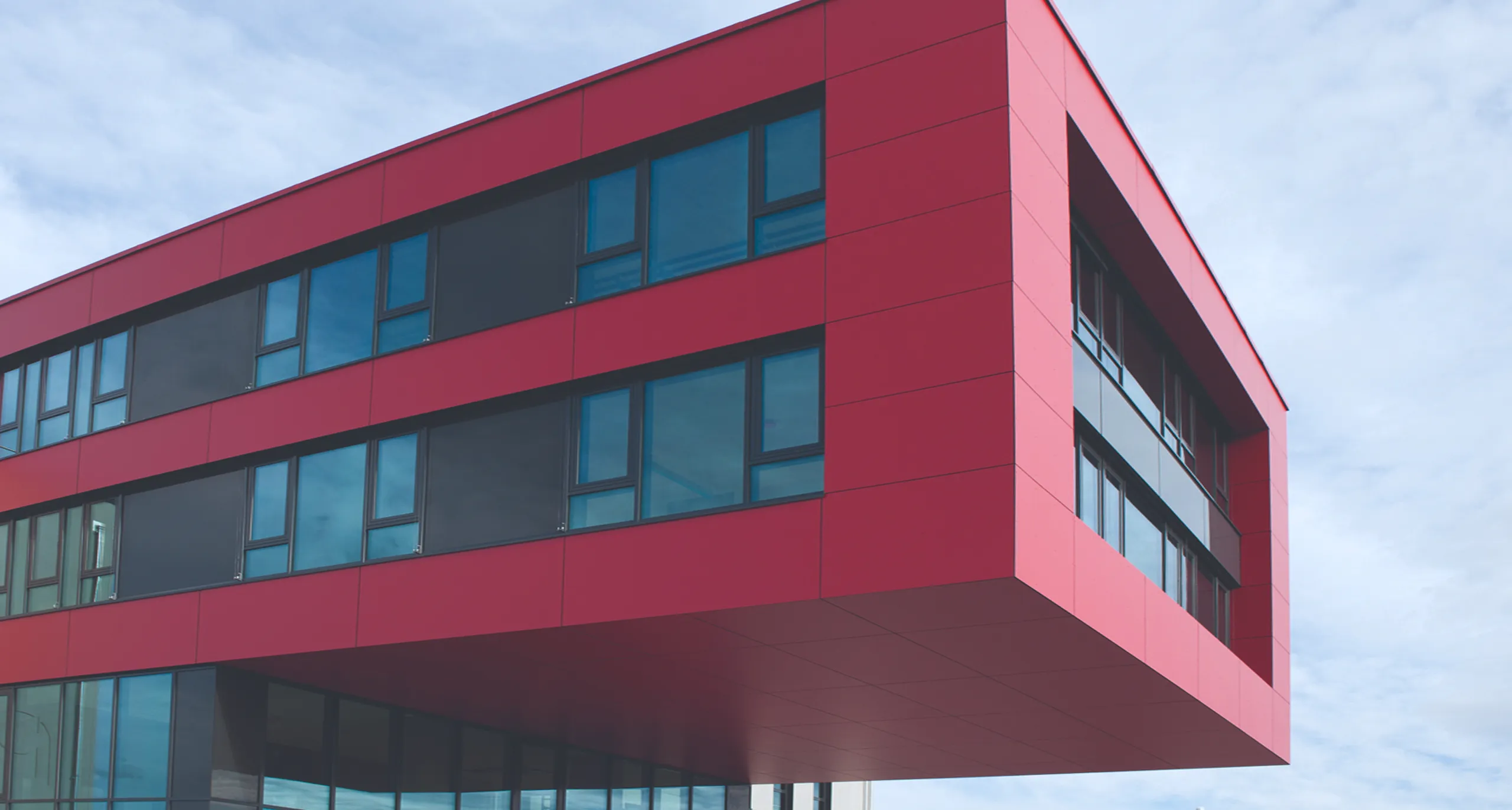 Modernes rotes Gebäude mit großen Fenstern und einem überstehenden oberen Stockwerk unter einem blauen Himmel.