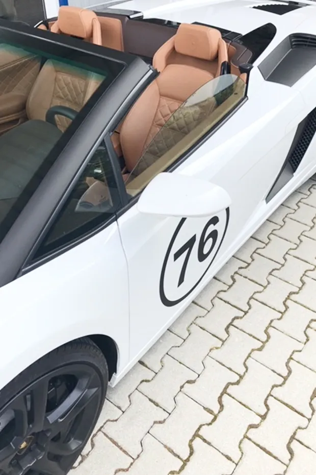 Weißer Sportwagen mit braunem Innenraum und der Nummer 76 auf der Seite, geparkt auf einem Pflastersteinboden.