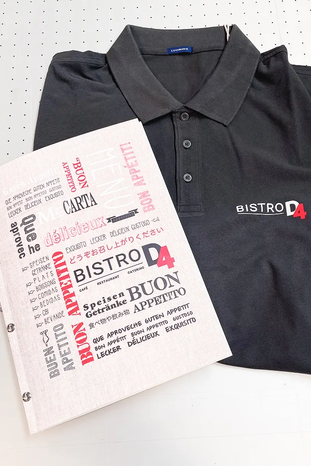 Schwarzes Polohemd mit dem Schriftzug "BISTRO D4" und einem Menüheft mit verschiedenen Speisenbezeichnungen.